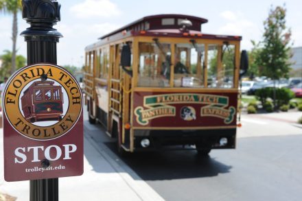 A Florida Tech trolley, slightly, pulls into a stop with the trolley stop sign prominent in the foreground.