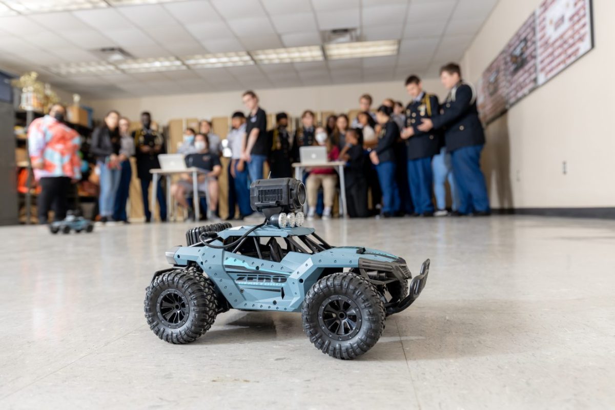 A remote-controlled car sits on the floor of a classroom with students in the background.